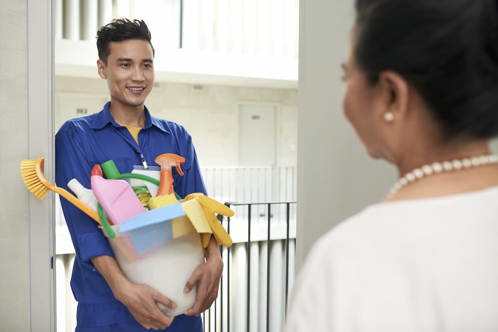 A friendly, uniformed male cleaner smiles at a customer while arriving at her door, holding a bucket filled with professional cleaning supplies.