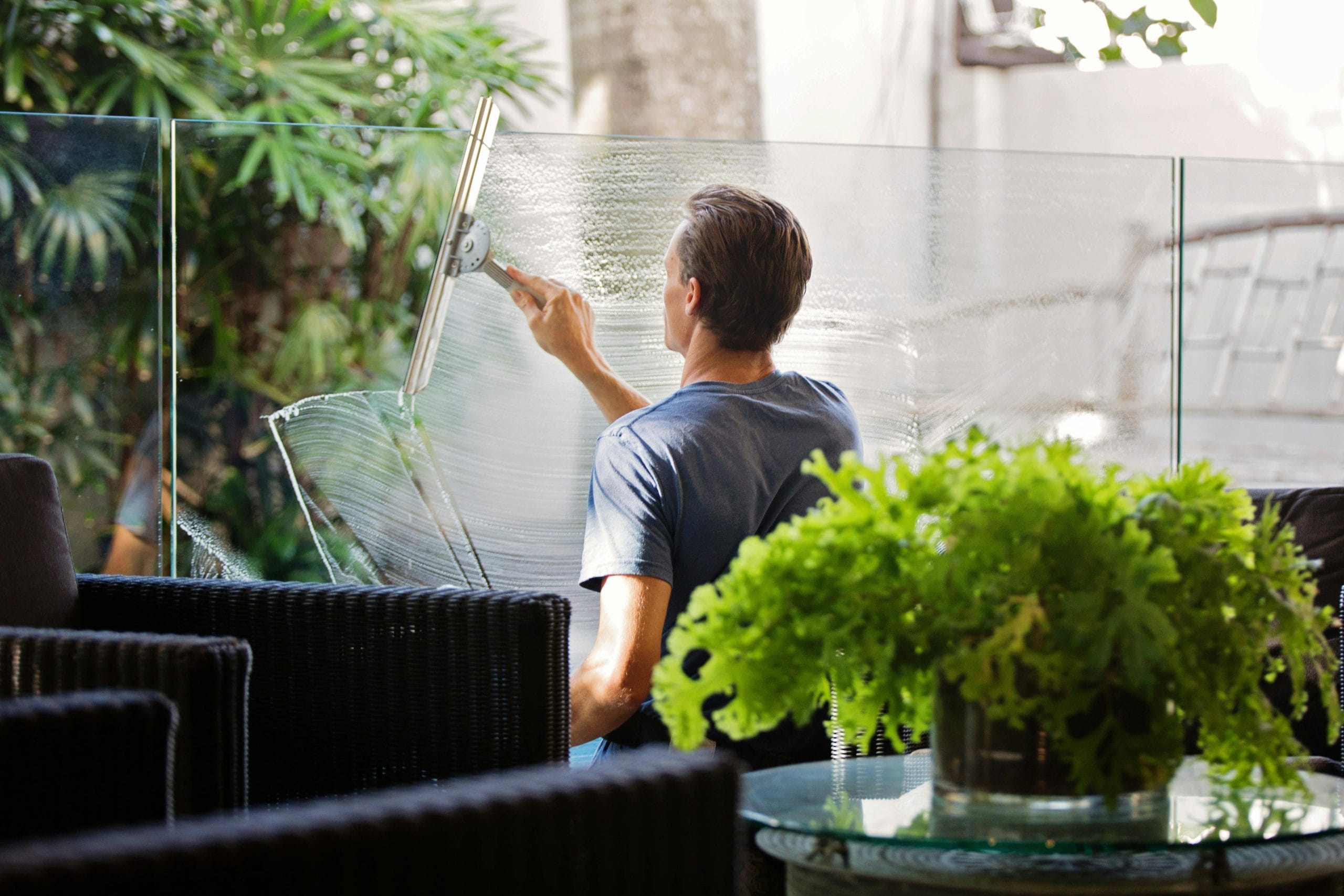A man seen from behind using a professional squeegee to clean a large glass partition on a patio, leaving a streak-free shine
