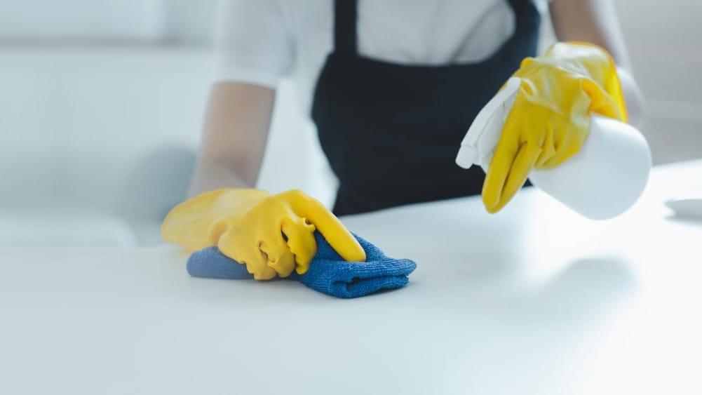 Surface) Close-up of a professional cleaner in yellow gloves sanitizing a white surface using a spray bottle and blue microfiber cloth.