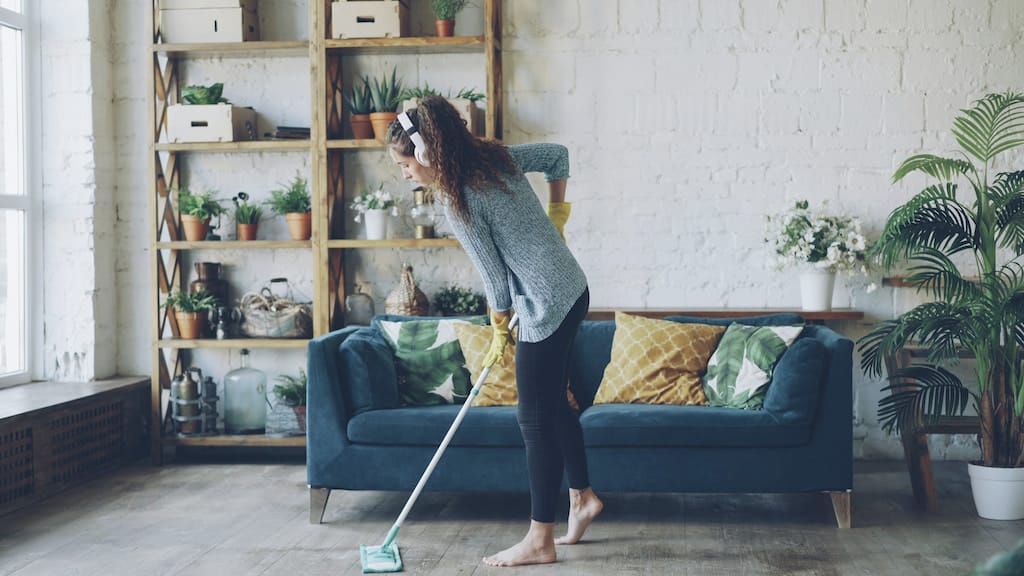 Woman wearing headphones and yellow gloves mopping a wooden floor in a modern living room.